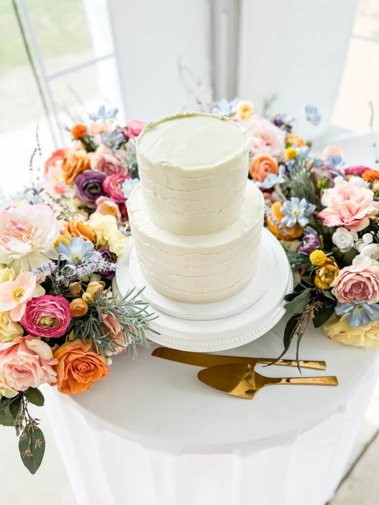 White two-tier cake surrounded by colorful fresh flowers